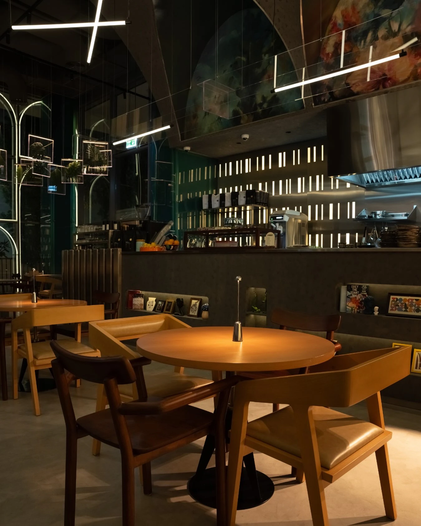 Arches traced in light and hanging frames holding plants beside the counter, a backlit slatted wall behind it, painted panels overhead and round wooden tables in the foreground