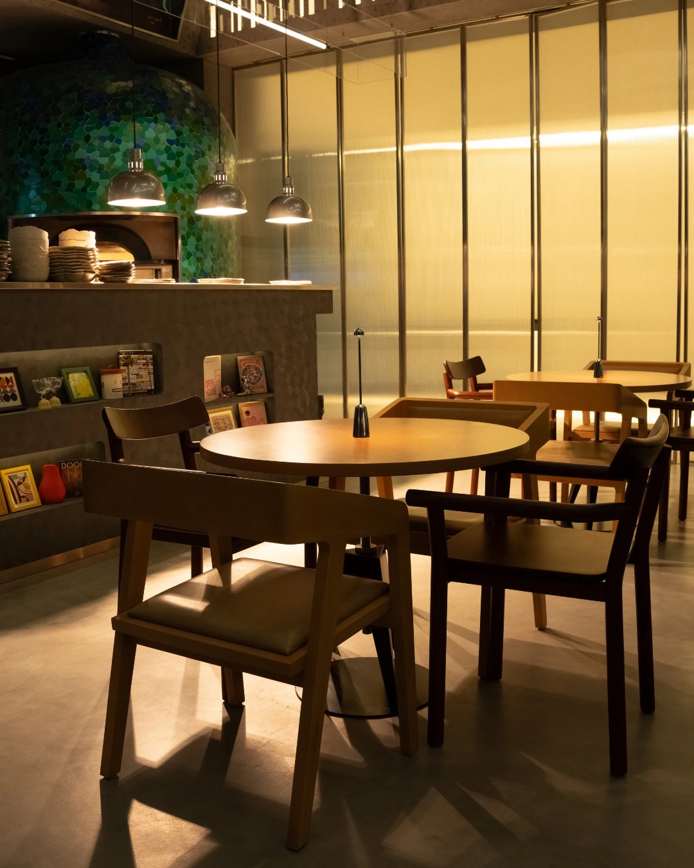 The dining room in warm low light: a green and blue mosaic-tiled pizza oven behind the counter, three pendant lamps, a glowing ribbed glass wall and round wooden tables with small table lamps