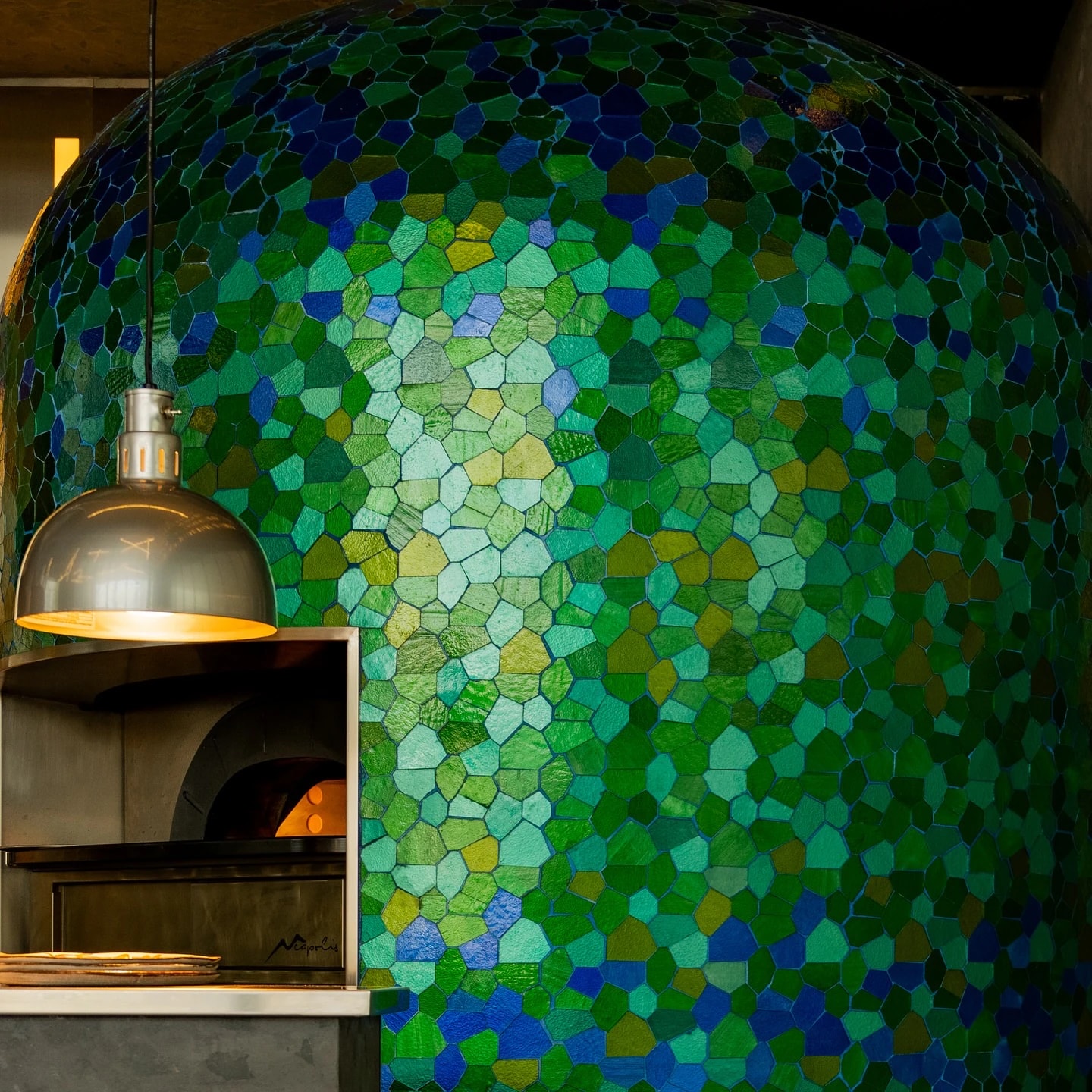 The pizza oven's dome tiled in green, blue and teal glass mosaic, a steel pendant lamp hanging over the oven mouth and a fire glowing inside