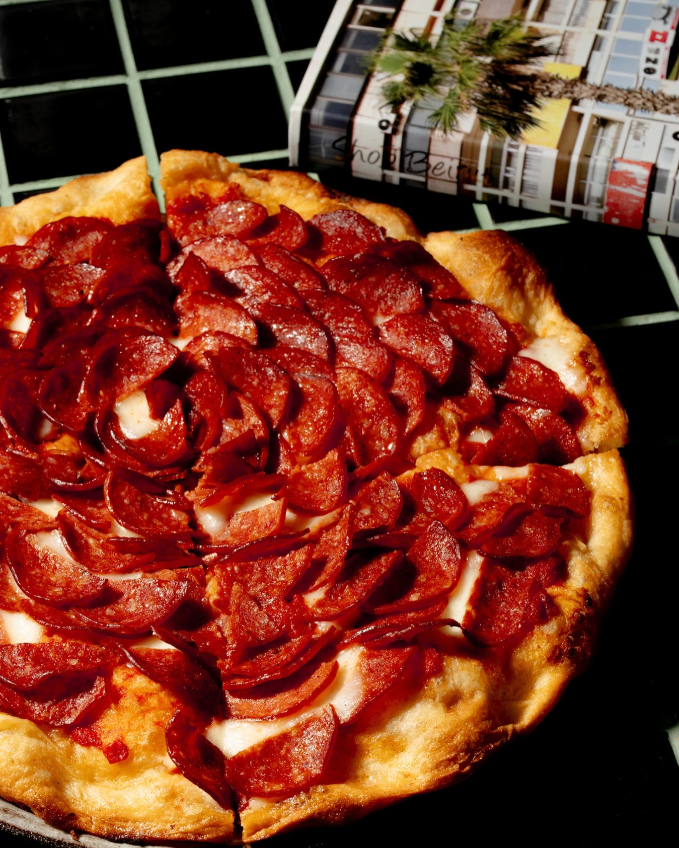 A pepperoni pizza covered edge to edge in cupped pepperoni, mozzarella showing through, a magazine behind it on the tiled table