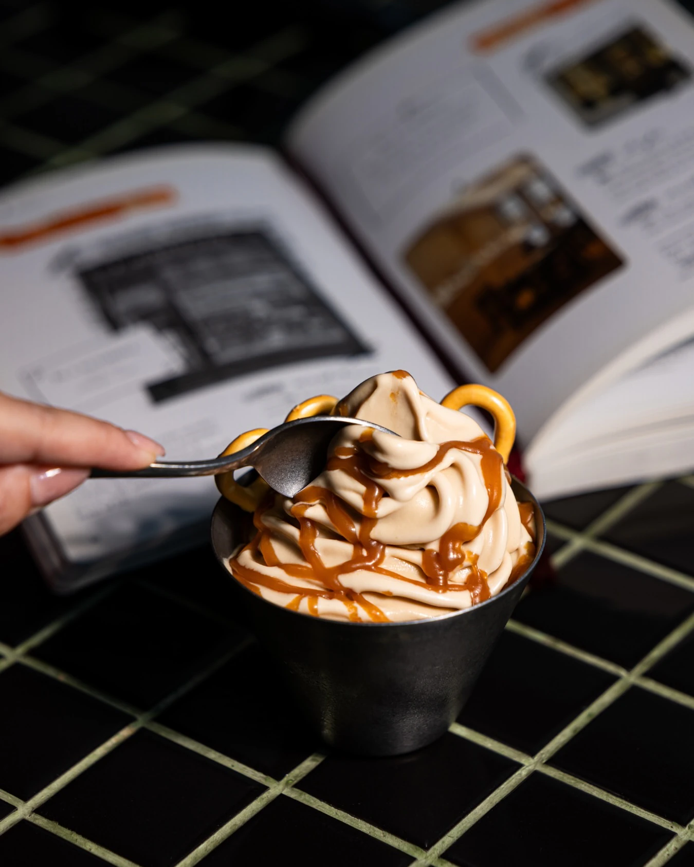 A swirl of pale soft serve drizzled with caramel in a pewter cup, pretzels tucked in and a spoon dipping in, an open book behind on a black tiled table