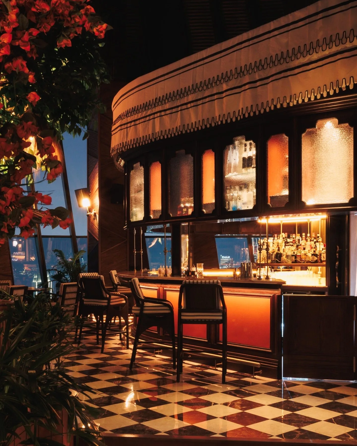 The bar at night: a striped, scalloped canopy over glowing amber glass cabinets and shelves of bottles, striped stools along an orange-lit counter, a checkerboard floor and red bougainvillea by the windows