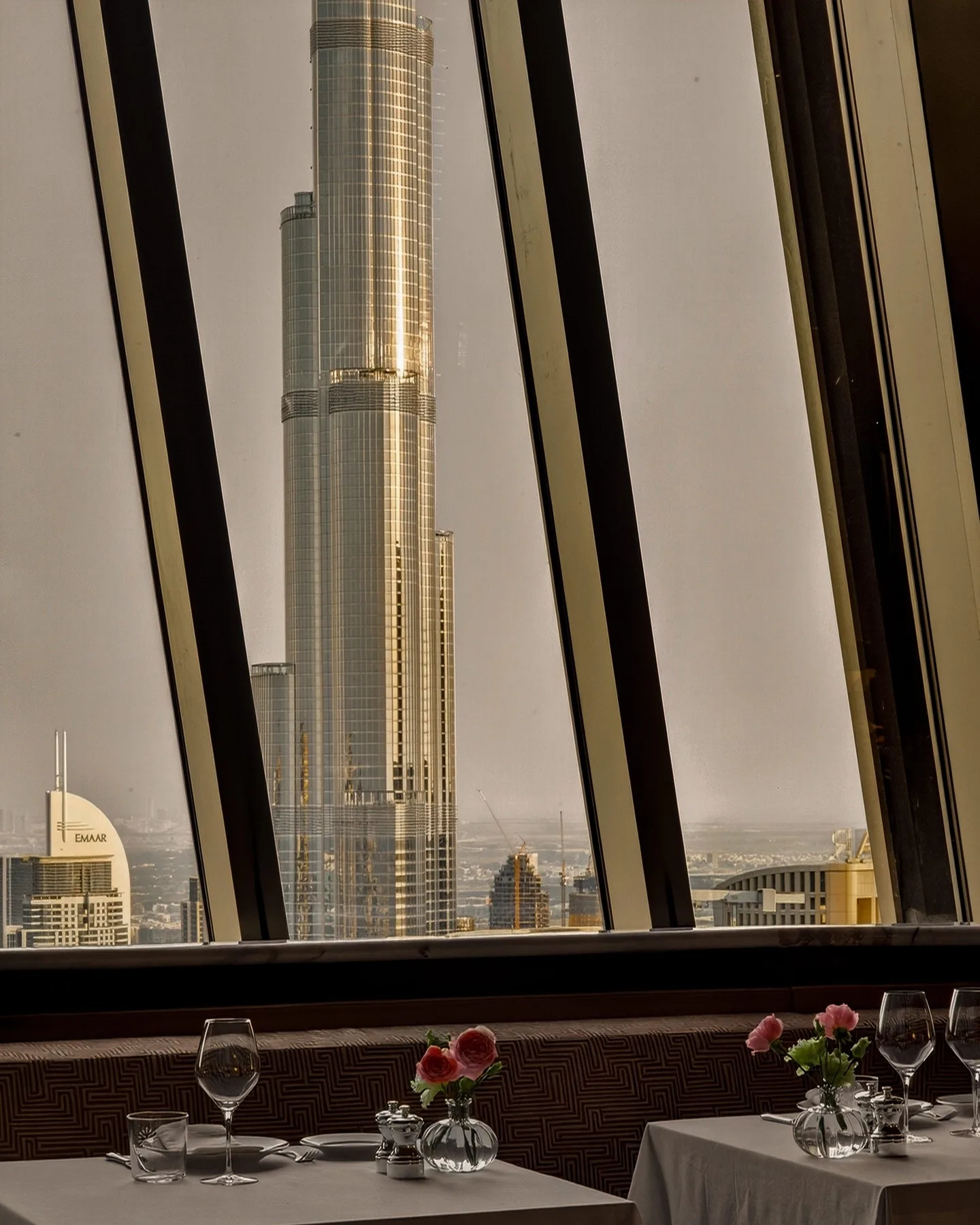The Burj Khalifa framed by tall slanted windows, an Emaar tower to its left, above two white-clothed tables with pink roses in glass vases, wine glasses and a patterned banquette
