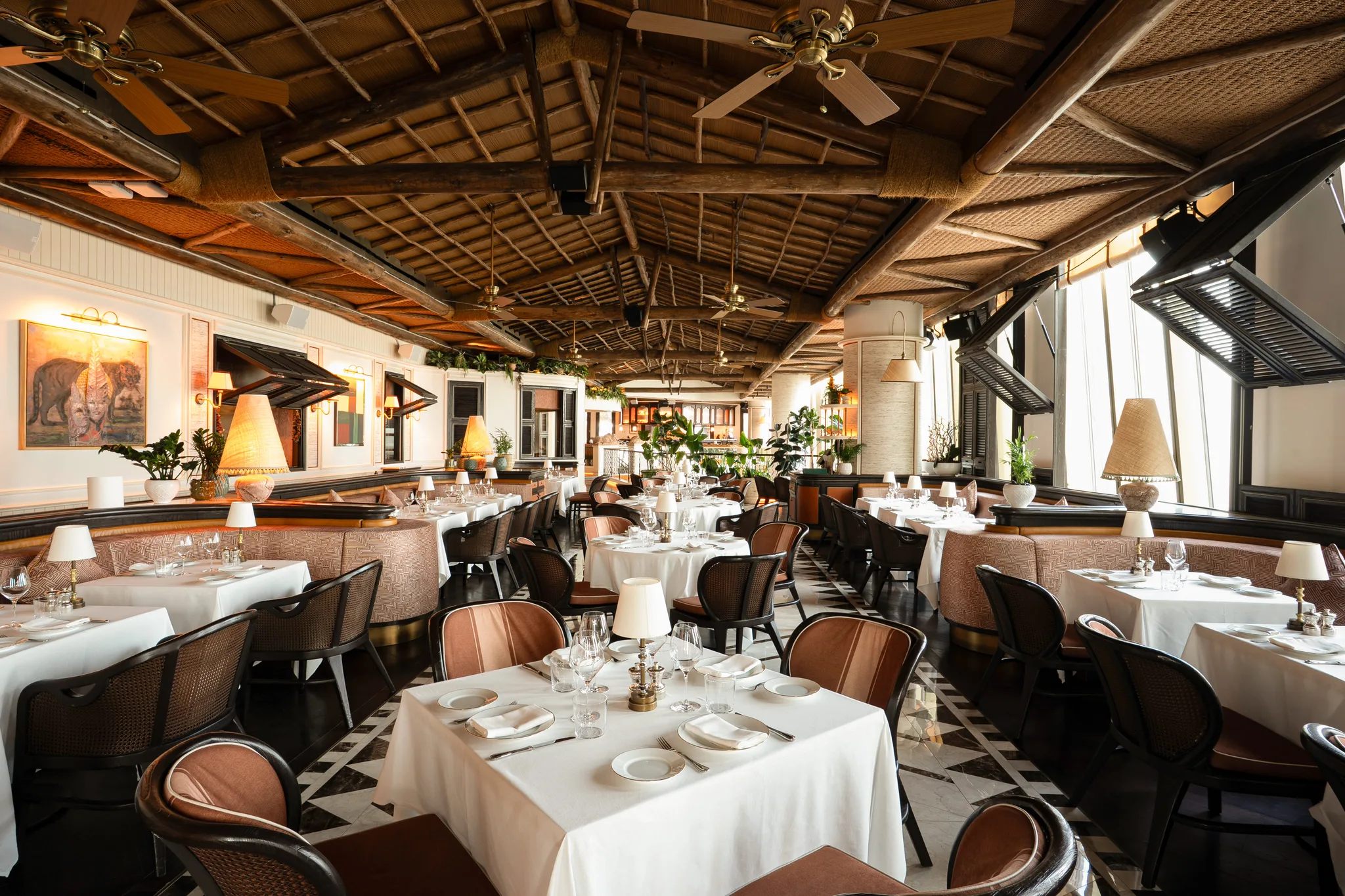 The dining room under a ceiling of timber beams and woven rattan with ceiling fans: white-clothed tables with small lamps, dark cane chairs with blush leather backs, banquettes, potted plants and shuttered windows on the right
