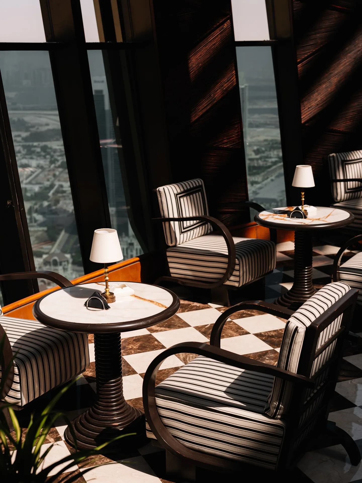 Striped black-and-white armchairs with curved dark-wood arms around round marble tables with small brass lamps, on a checkerboard marble floor, sunlight falling through tall windows over the city far below