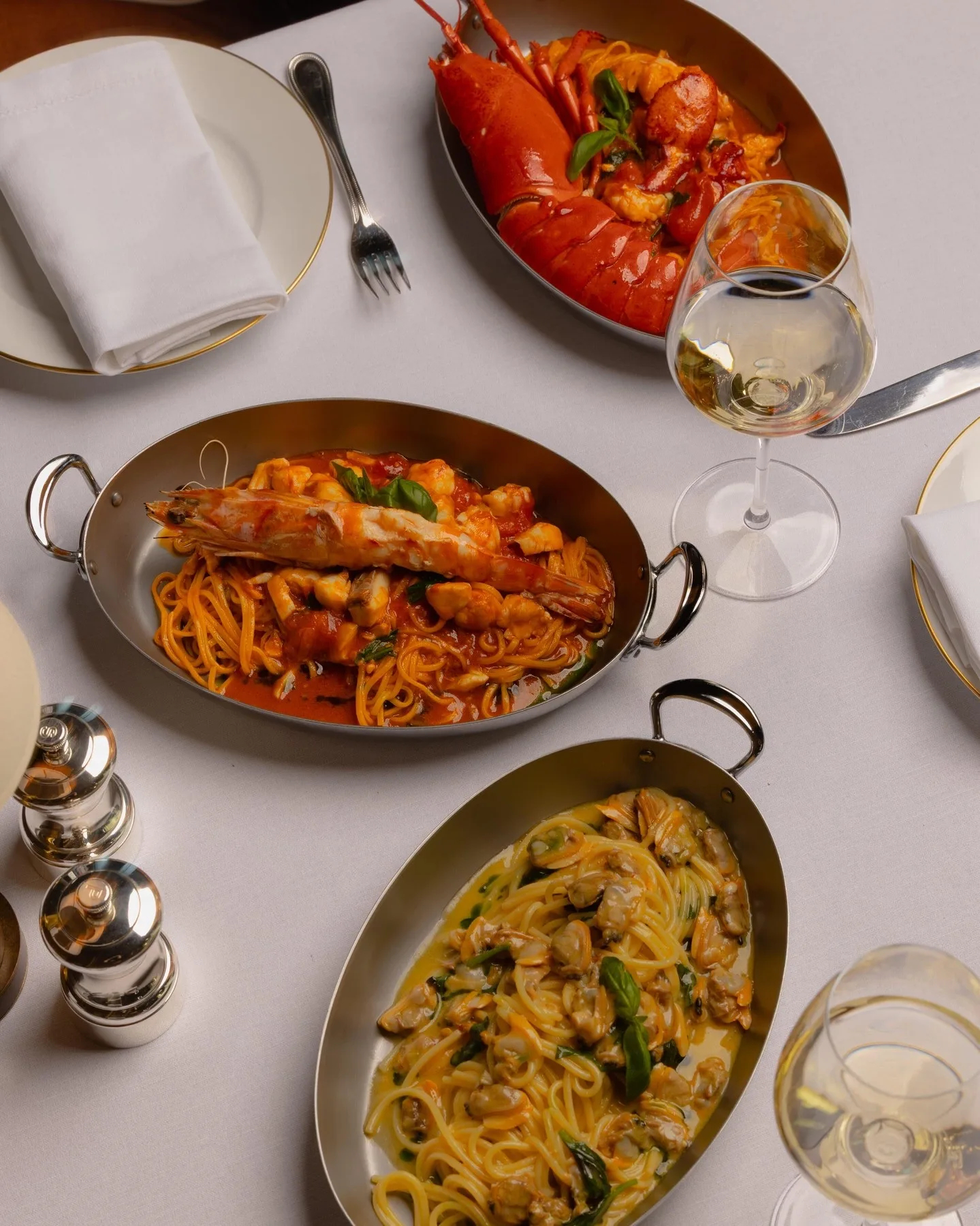 Three pastas in oval steel pans on a white tablecloth: spaghetti with a halved red lobster in tomato sauce, spaghetti in tomato sauce with seafood under a whole prawn, and spaghetti with clams and basil, beside glasses of white wine and silver salt and pepper mills