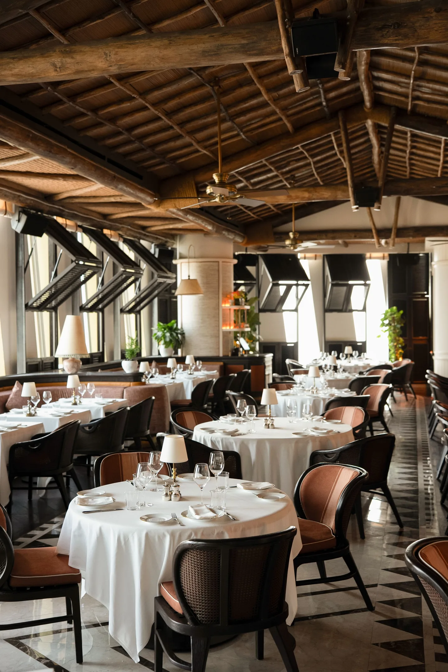 Round tables in long white cloths with small lamps and dark cane chairs with tan leather backs, under a timber-beamed ceiling with fans, on a patterned marble floor
