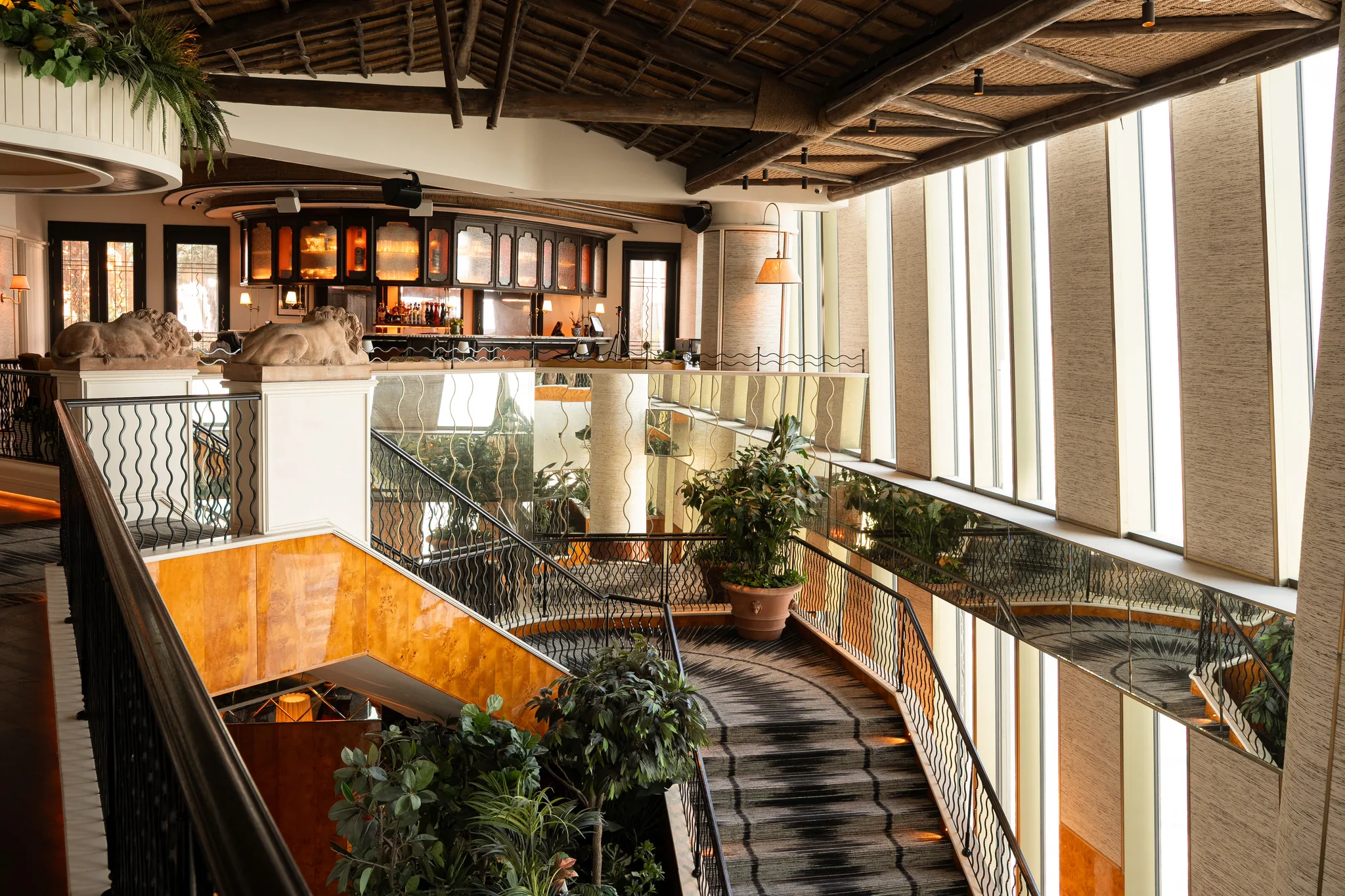 A curving staircase with wavy black iron railings between two floors, a pair of stone lions on white plinths at the top, the bar's glowing cabinets beyond, potted plants and tall windows with woven blinds