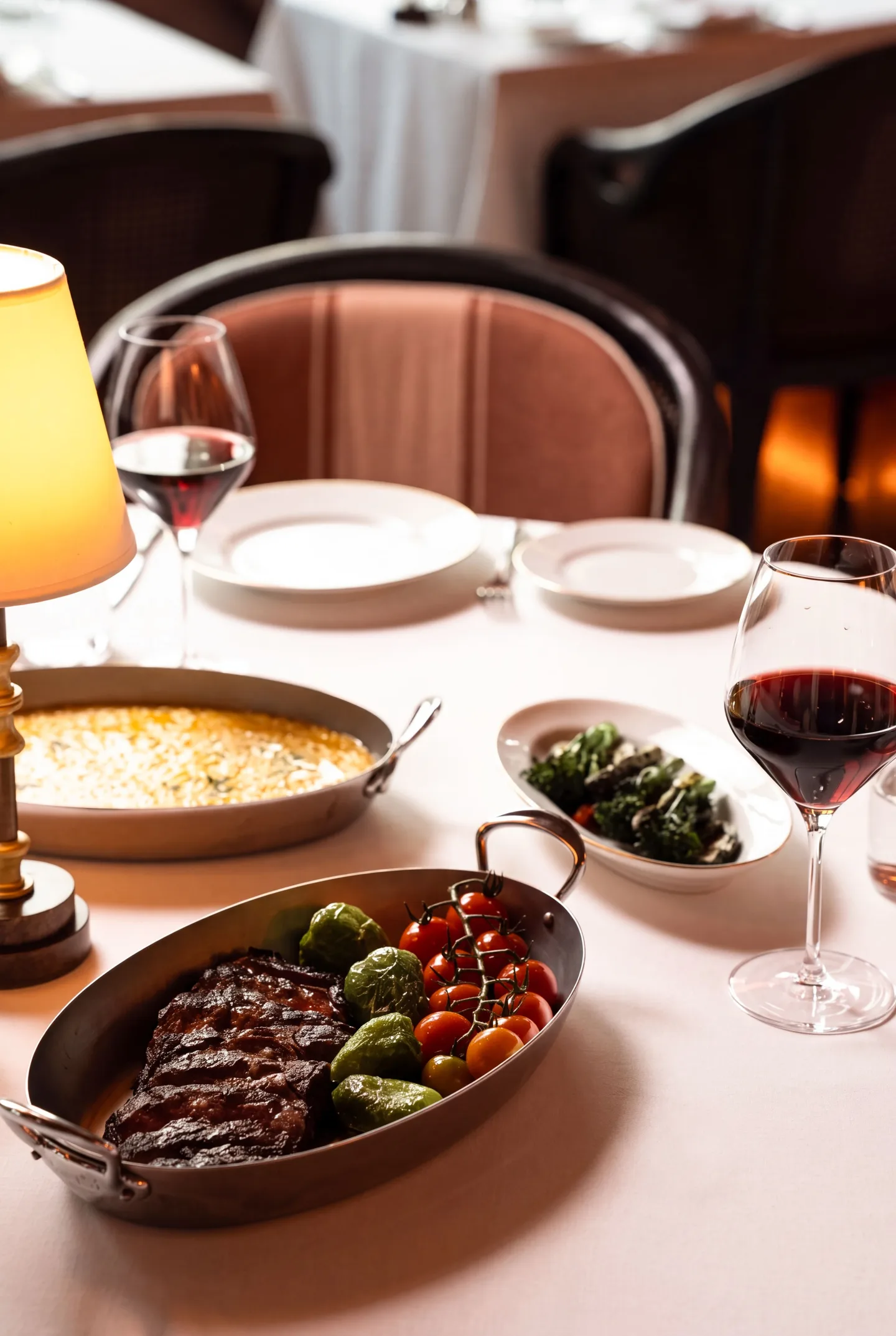 A grilled steak in an oval steel pan with Brussels sprouts and cherry tomatoes on the vine, beside a gratin, a dish of greens and two glasses of red wine under a table lamp