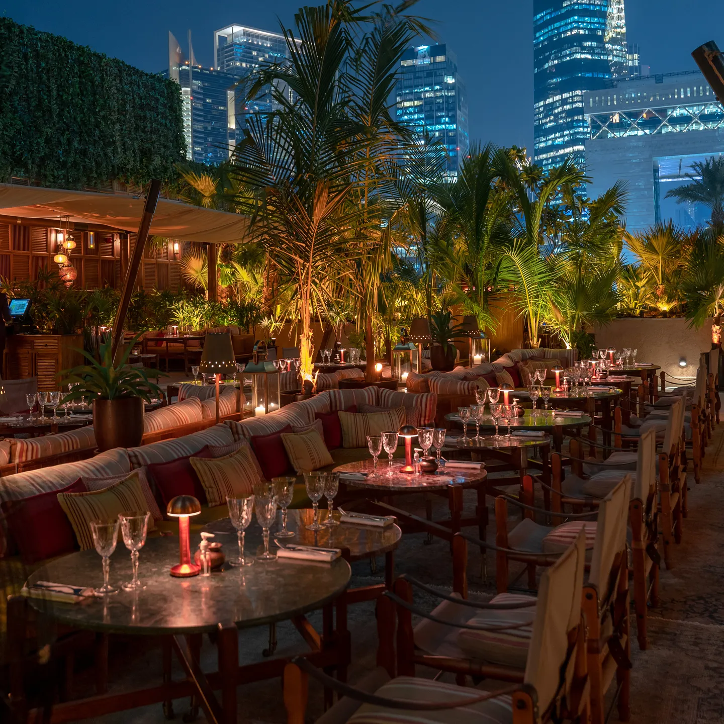 Lamplit tables and striped sofas on the terrace at night, towers rising behind