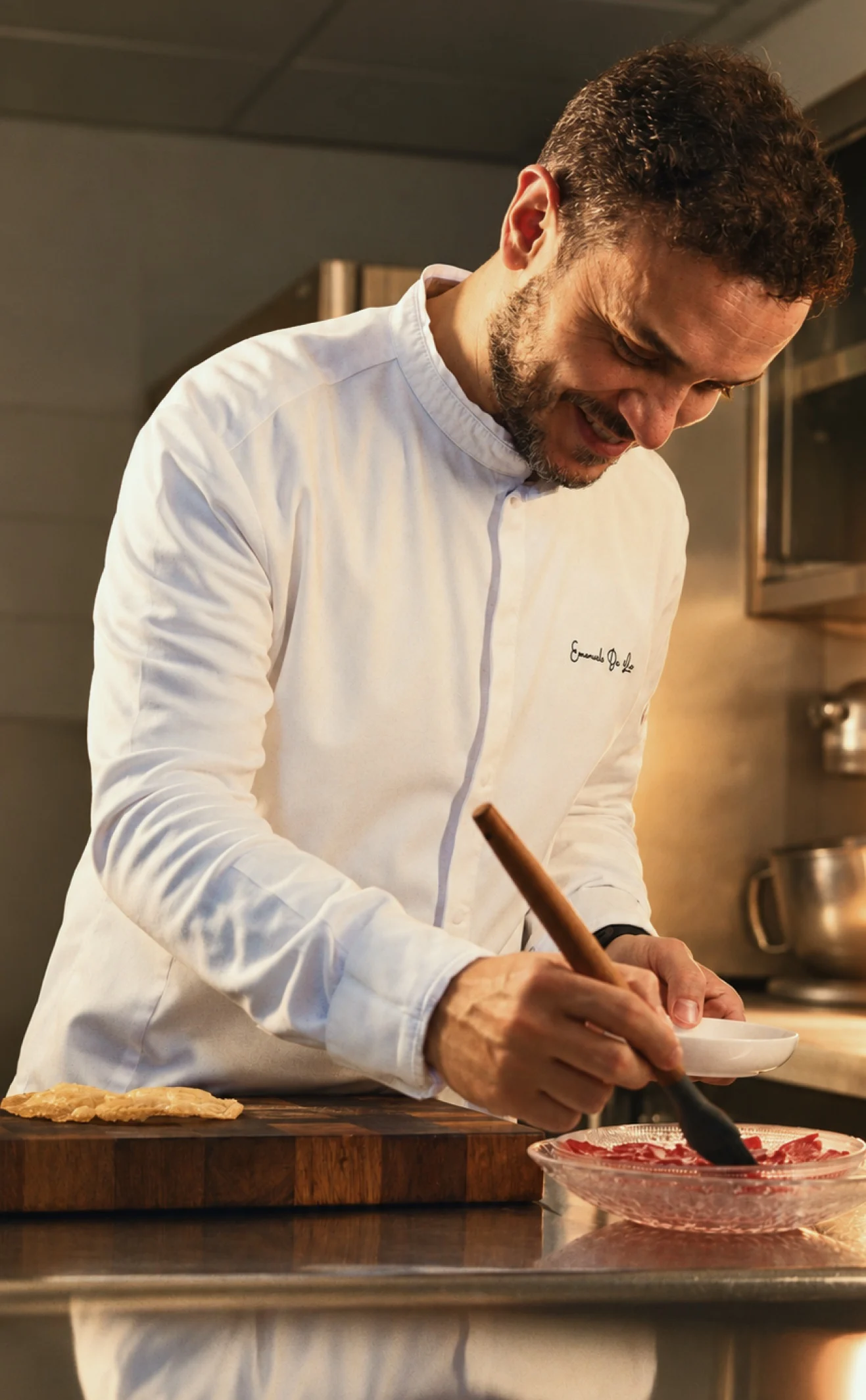 Chef Emanuele De Leo in chef's whites, smiling over his work at a steel counter, a wooden-handled brush in one hand and a small white bowl in the other