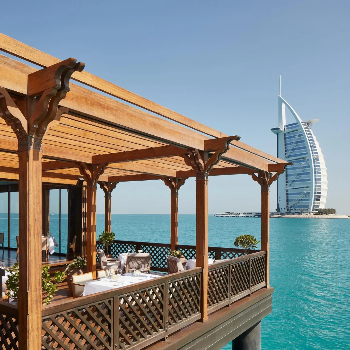 The terrace under a timber pergola with lattice railings, white-clothed tables over the turquoise water and the Burj Al Arab across the bay