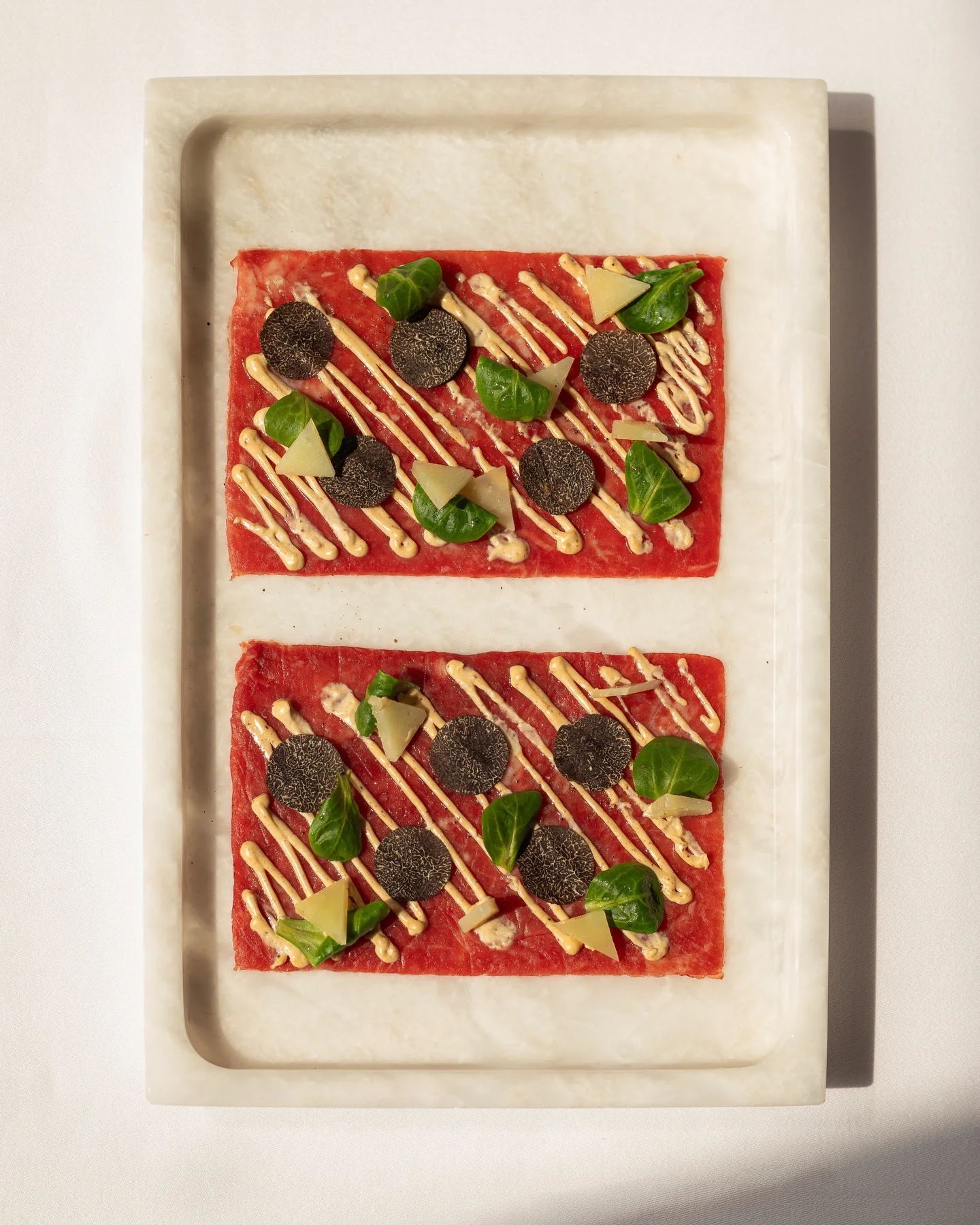 Overhead view of two rectangles of carpaccio on a marble tray, topped with slices of black truffle, a drizzled sauce, small green leaves and shavings of cheese