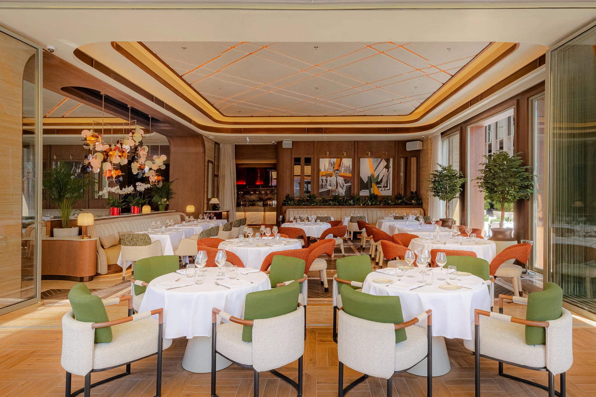 The dining room by day: round white-clothed tables with cream and green armchairs in front, orange chairs beyond, glass-disc chandeliers, a gold-trimmed ceiling and paintings on a wood-panelled wall