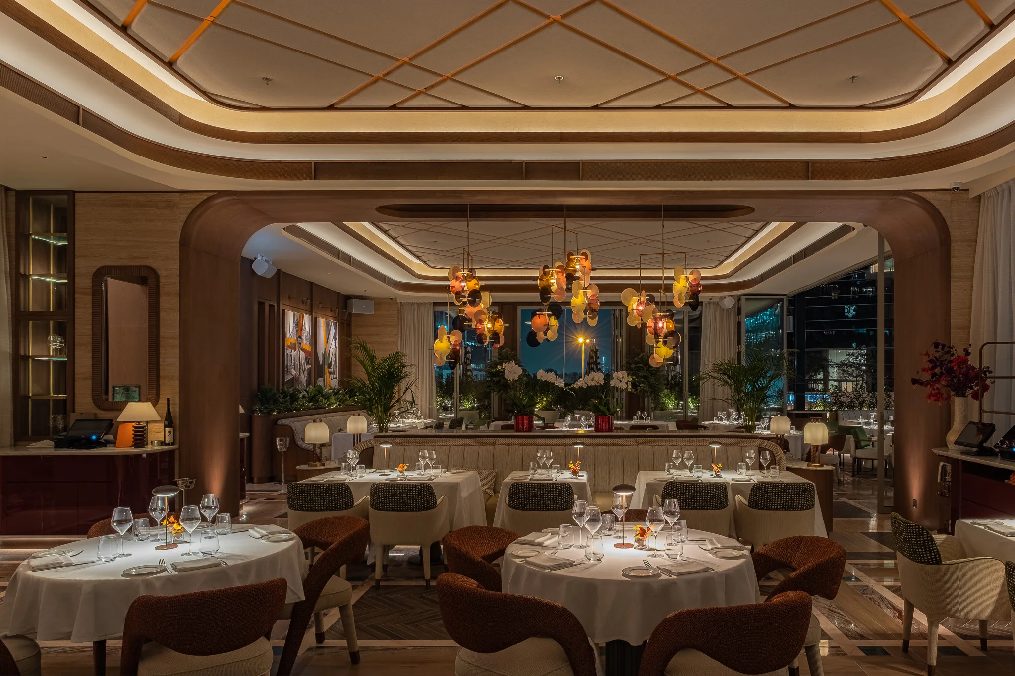 The dining room at night: an arched opening framing clusters of amber glass-disc chandeliers, white-clothed tables with rust and patterned chairs, a long banquette and the lit towers of DIFC through the windows
