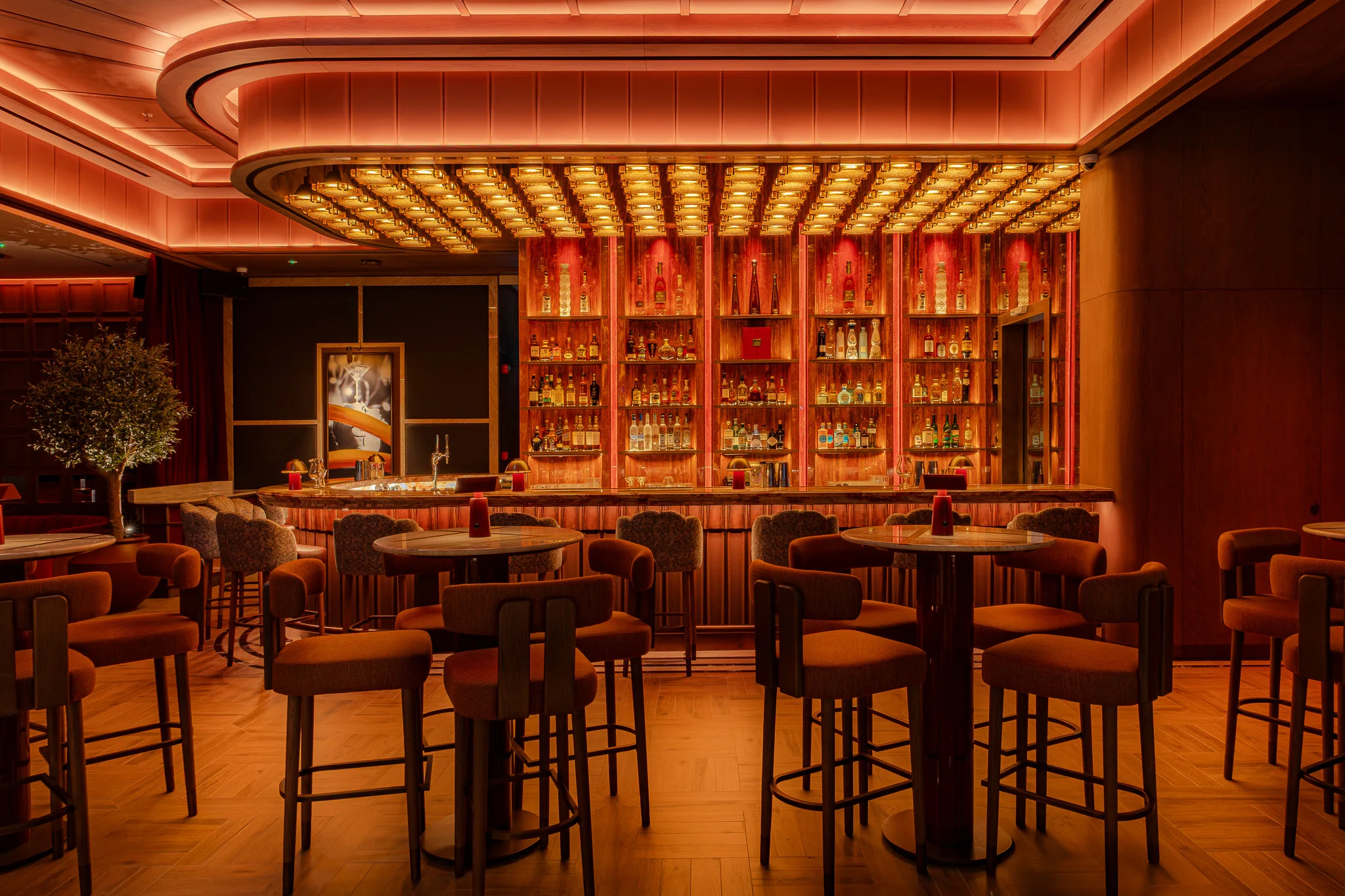 A bar lit red: backlit shelves of bottles under a ceiling grid of small lights, high tables and rust velvet bar stools on a herringbone floor