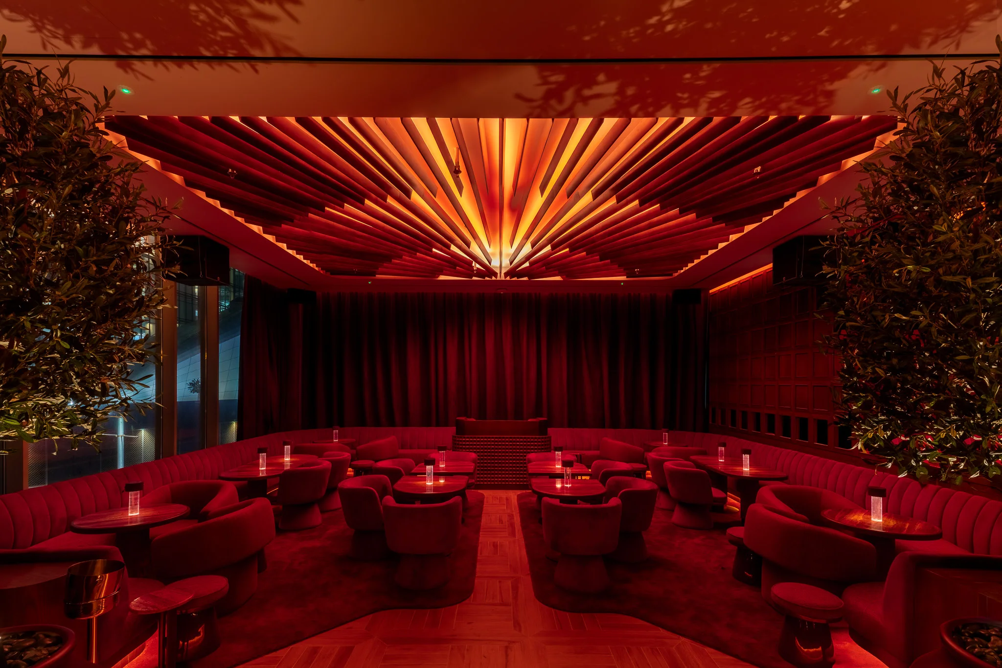 A lounge in red: velvet booths and armchairs around low tables with glowing candles, a drawn red curtain at the far end under a fanned, lit ceiling, olive trees on either side