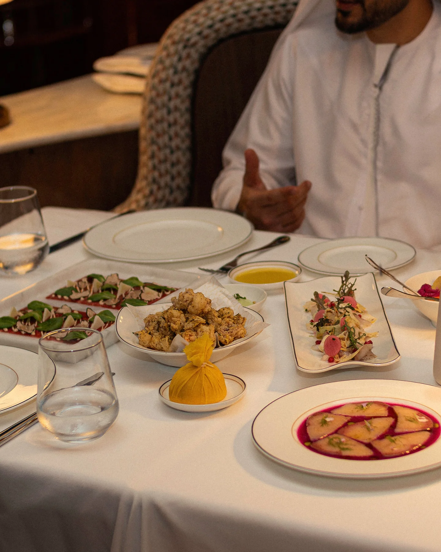 Shared plates on a white tablecloth: carpaccio with green leaves and shavings on a white tray, a bowl of fritti on paper, a lemon wrapped in yellow muslin, raw fish with radish and herbs, and sliced raw fish in a pink sauce, a guest in a kandura mid-conversation behind