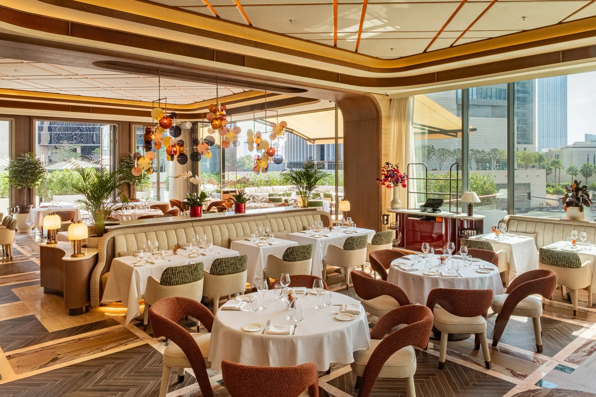The dining room in daylight: rust and cream chairs around white-clothed tables, glass-disc chandeliers and a long banquette, and through the glass walls the terrace's sofas, palms and the DIFC towers