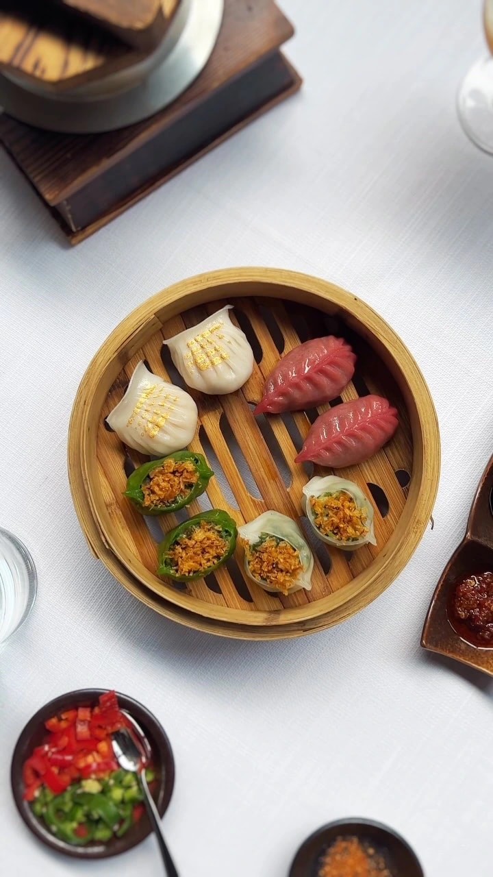 Overhead view of a bamboo steamer holding eight pieces of dim sum: white dumplings flecked with gold, red leaf-shaped dumplings, stuffed green peppers and open dumplings with a crisp topping, chili dishes beside it