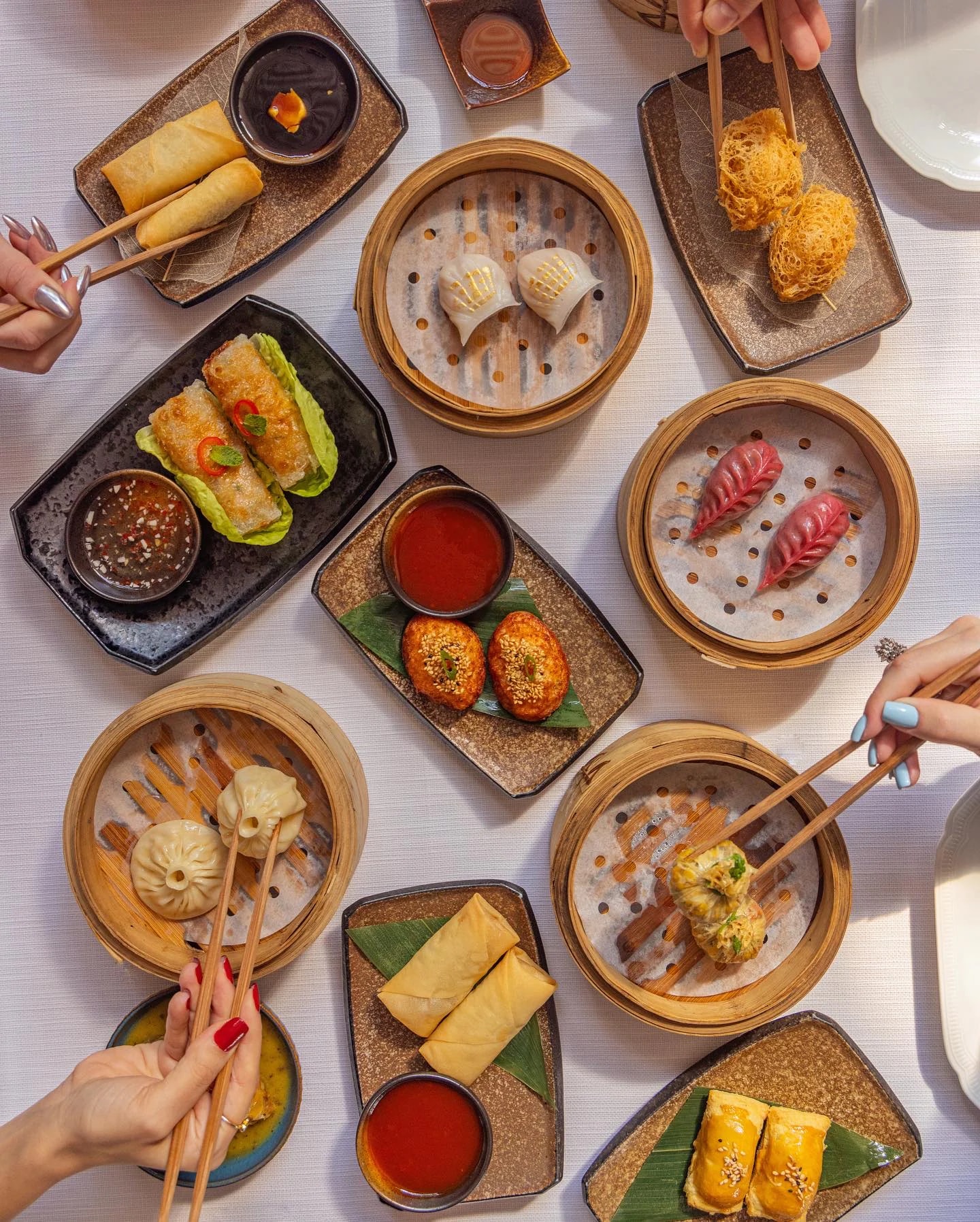 Overhead view of dim sum on a white tablecloth: bamboo steamers of dumplings, spring rolls, golden crisp prawns and sesame-topped cakes with dipping sauces, hands reaching in with chopsticks