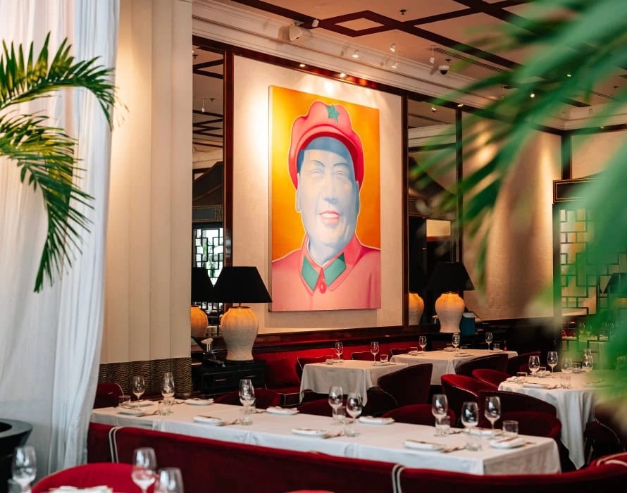 The dining room: a large orange and pink pop-art portrait above red velvet banquettes, black-shaded lamps and white-clothed tables, palm fronds in the foreground