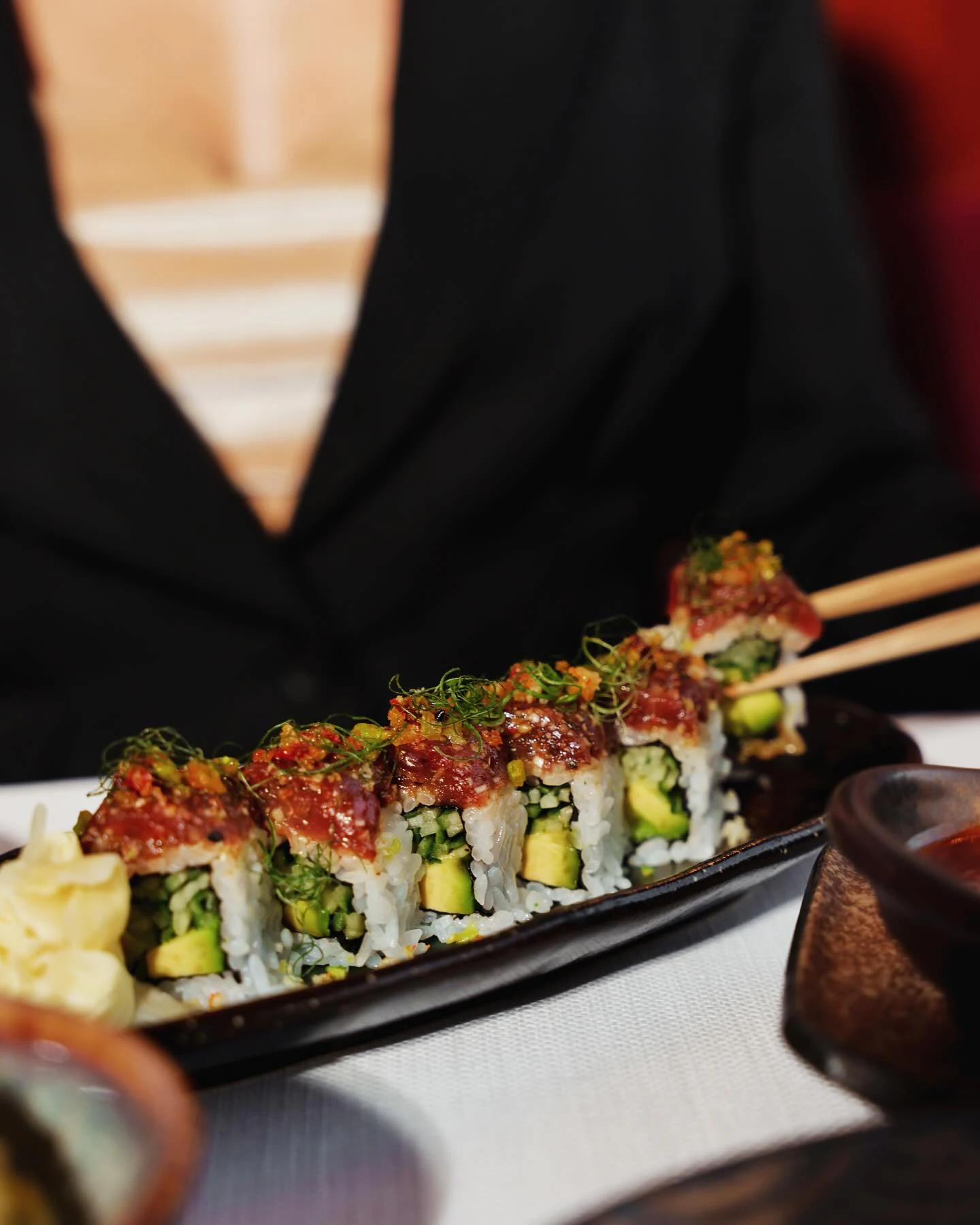 A maki roll with a red topping and avocado on a dark plate, one piece lifted with chopsticks