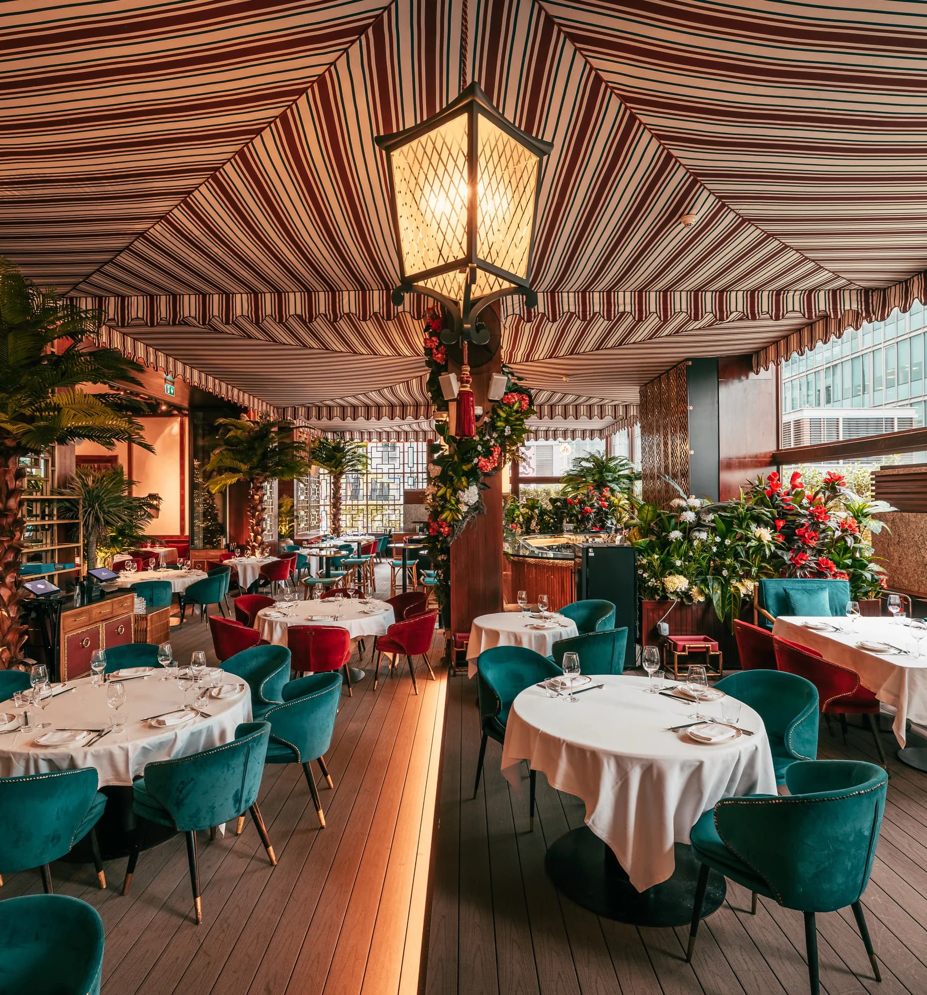 The terrace under a striped canopy: a lattice lantern hanging over a flower-wrapped pillar, palms, and round white-clothed tables with teal and red velvet chairs, glass towers through the windows