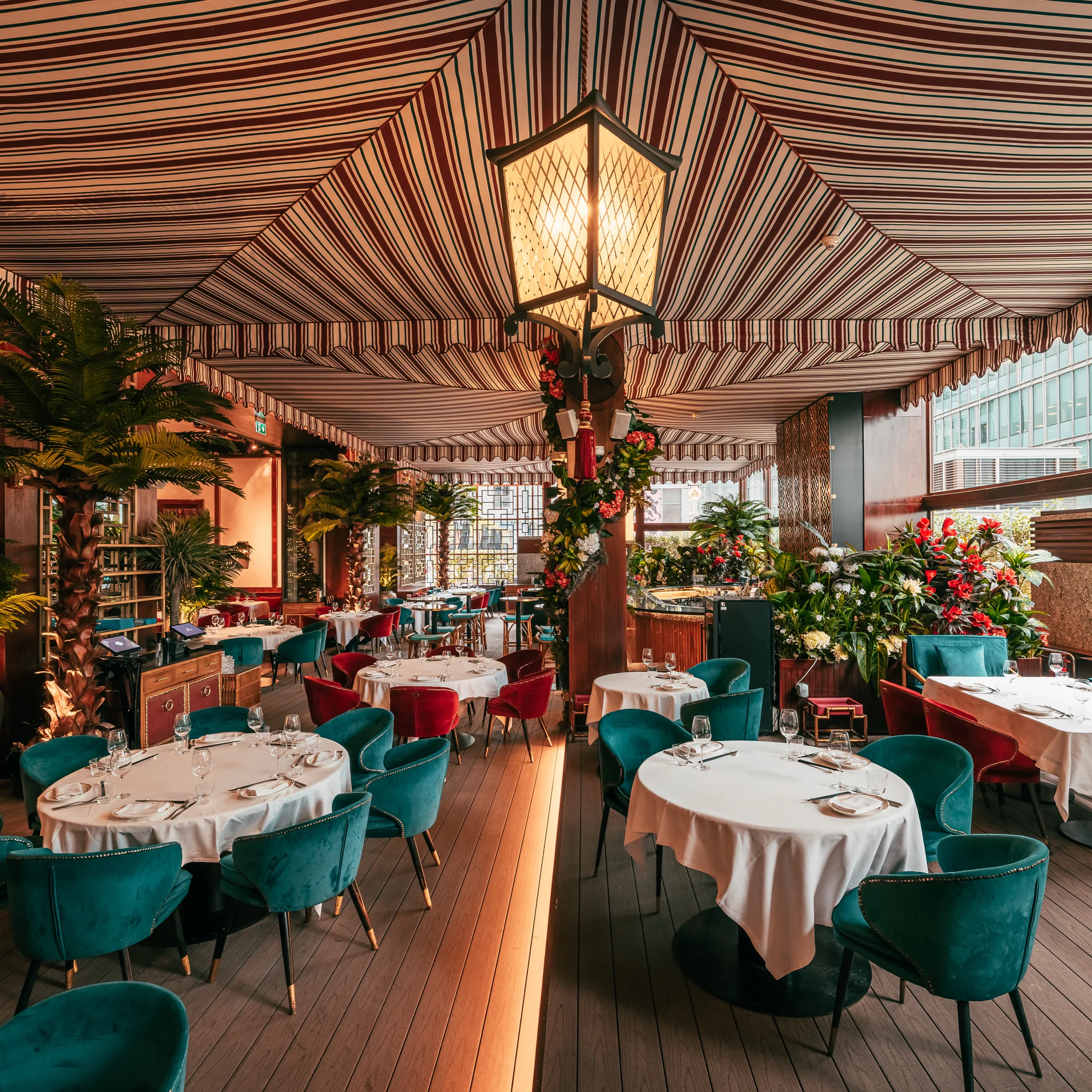 The terrace under a striped canopy: a lattice lantern hanging over a flower-wrapped pillar, palms, and round white-clothed tables with teal and red velvet chairs, glass towers through the windows