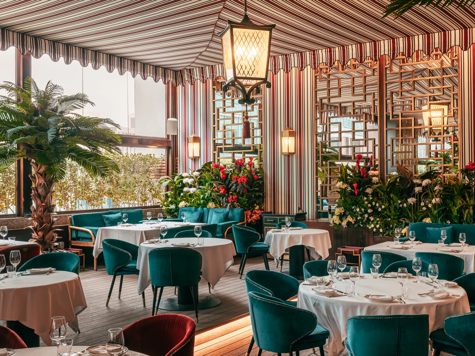 The terrace under a striped canopy: a hanging lantern, gold lattice screens, banks of red and white flowers, a palm and white-clothed tables with teal velvet chairs