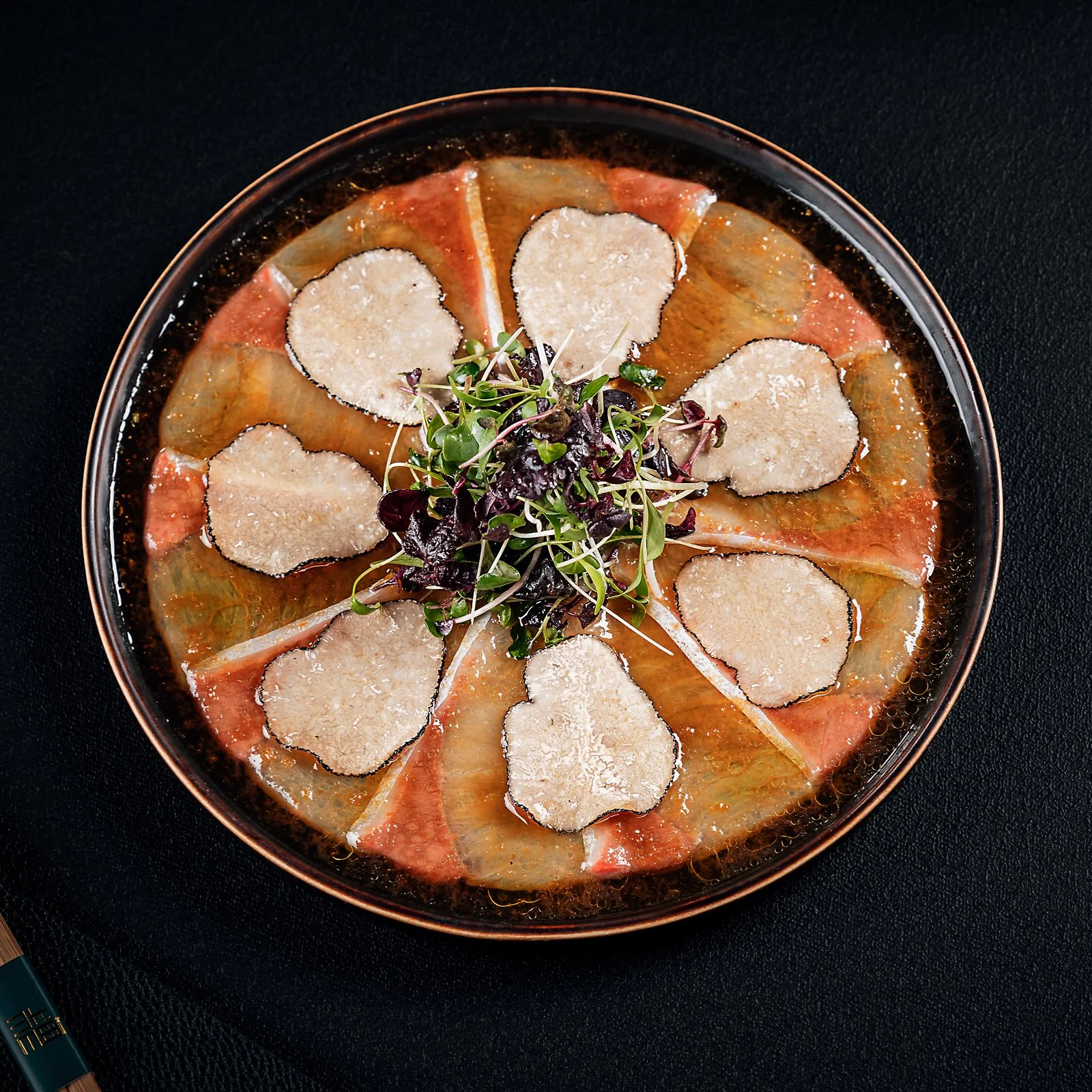 Slices of raw fish fanned out on a dark plate under shaved truffle, a tangle of microgreens at the centre