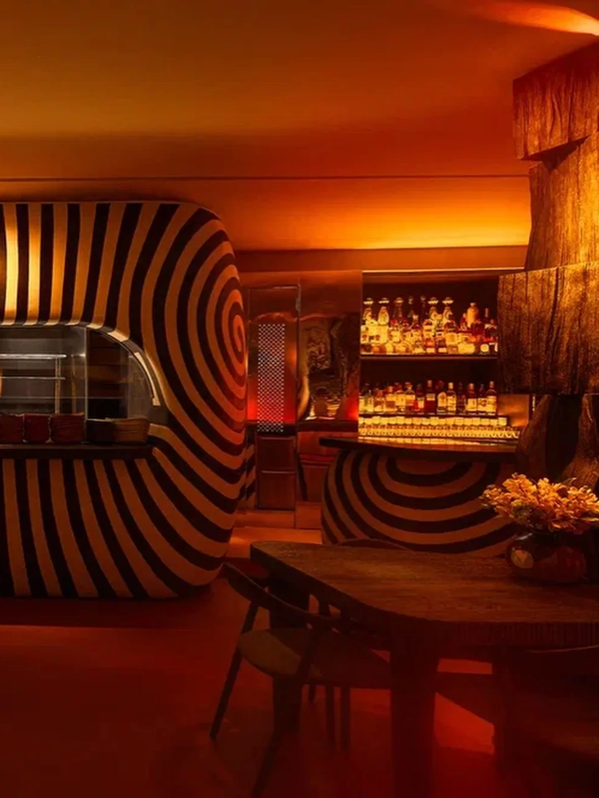 The bar in amber light: a black and white spiral-striped wall around the kitchen pass, bottles on lit shelves and a charred wood sculpture