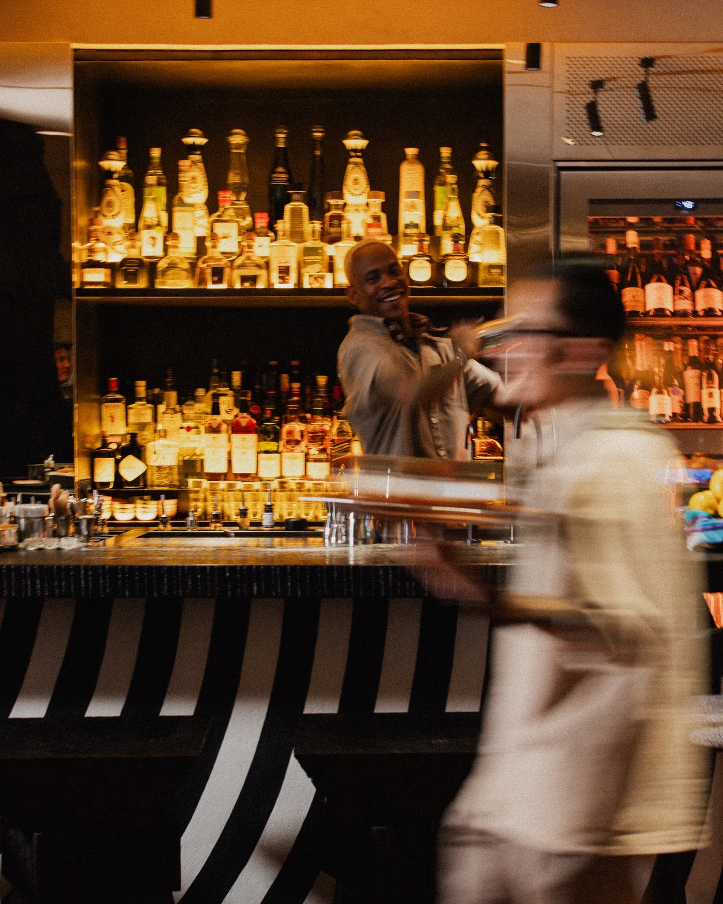 A bartender smiling behind the bar, bottles lit on the shelves behind him, a server blurred in motion in the foreground