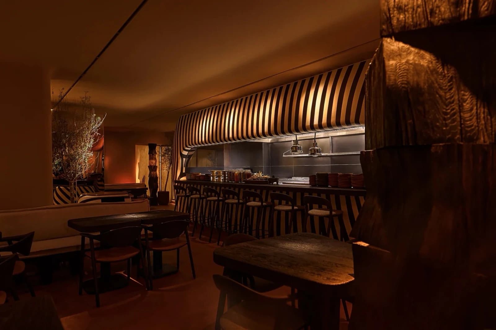 The dining room in low amber light: dark wooden tables, stools at the open kitchen's counter beneath a black and white striped canopy, and a charred wood sculpture in the foreground