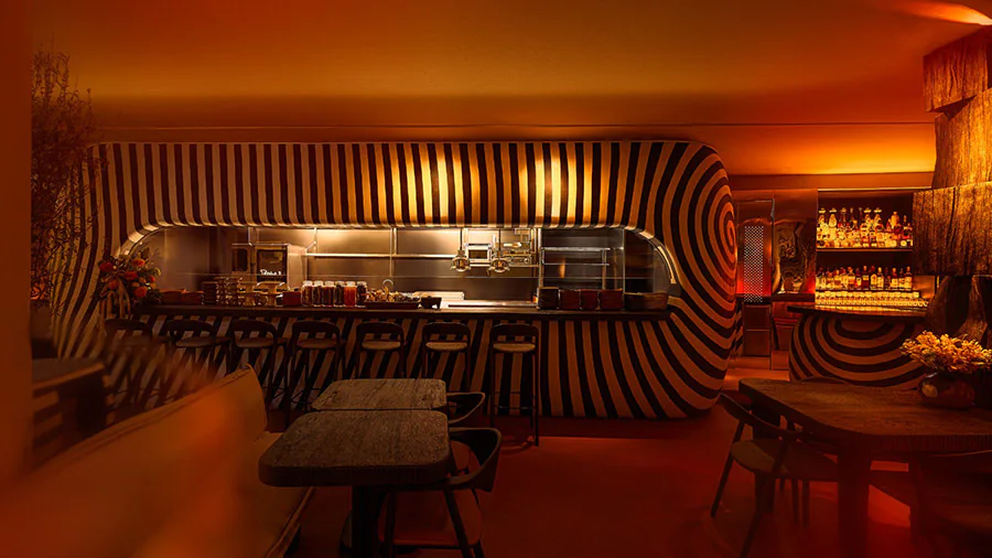 The dining room in amber light, the open kitchen wrapped in black and white stripes, stools along its counter and the bar to the right