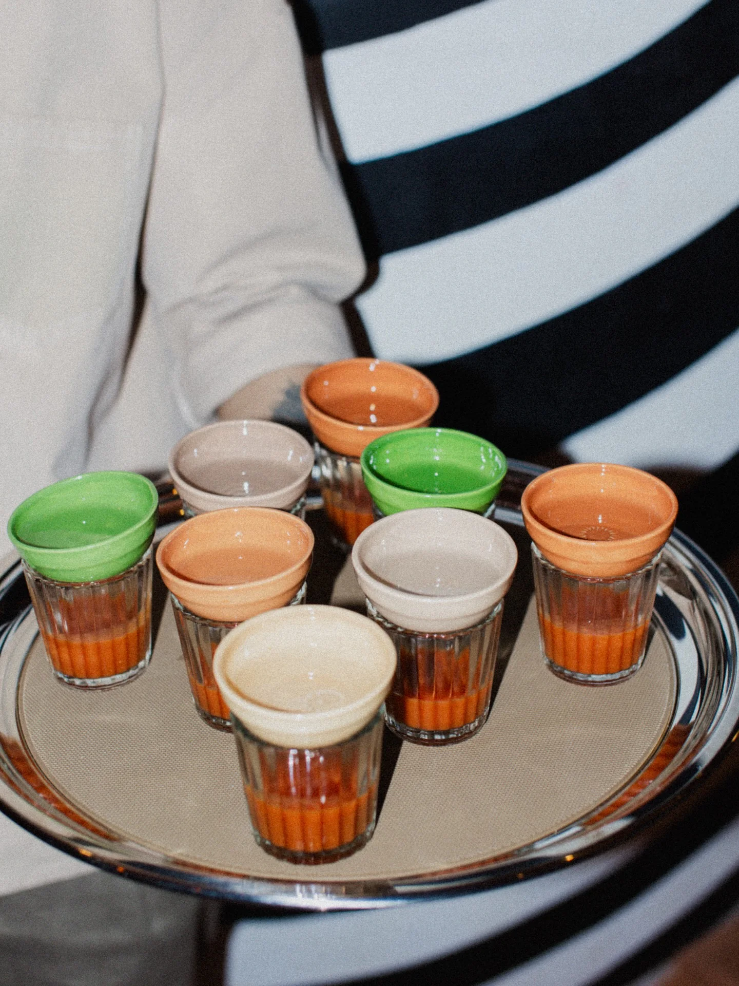 A server holding a round tray of small glasses of an orange drink, each capped with a glazed green, orange or cream cup