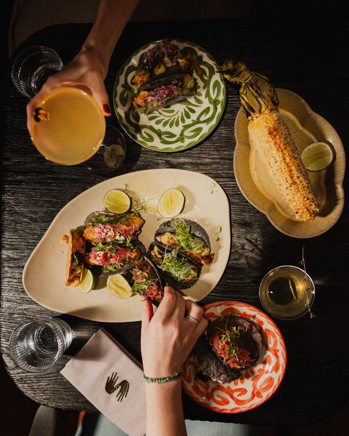 Overhead view of a dark wooden table: blue corn tacos and lime on a cream platter, grilled street corn, cocktails and hands reaching across