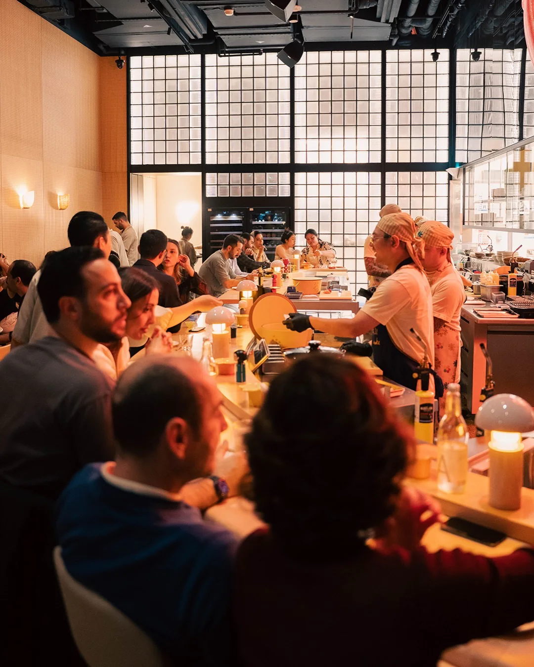 A chef in a bandana and black gloves working at the long counter while diners sit along it, a glass-block wall behind
