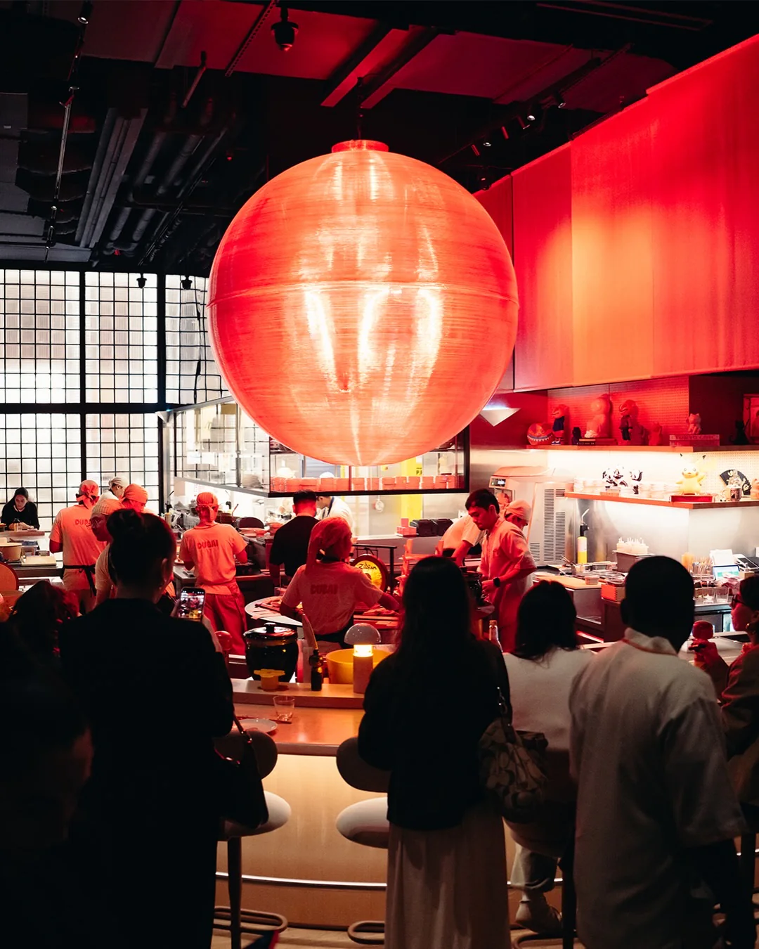 The red lantern glowing over the counter at night, chefs at work and a crowd gathered around the stools