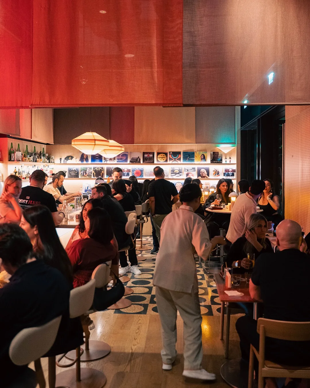 A busy evening in the dining room, red fabric panels overhead, bottles behind the bar and record sleeves lit along the back wall