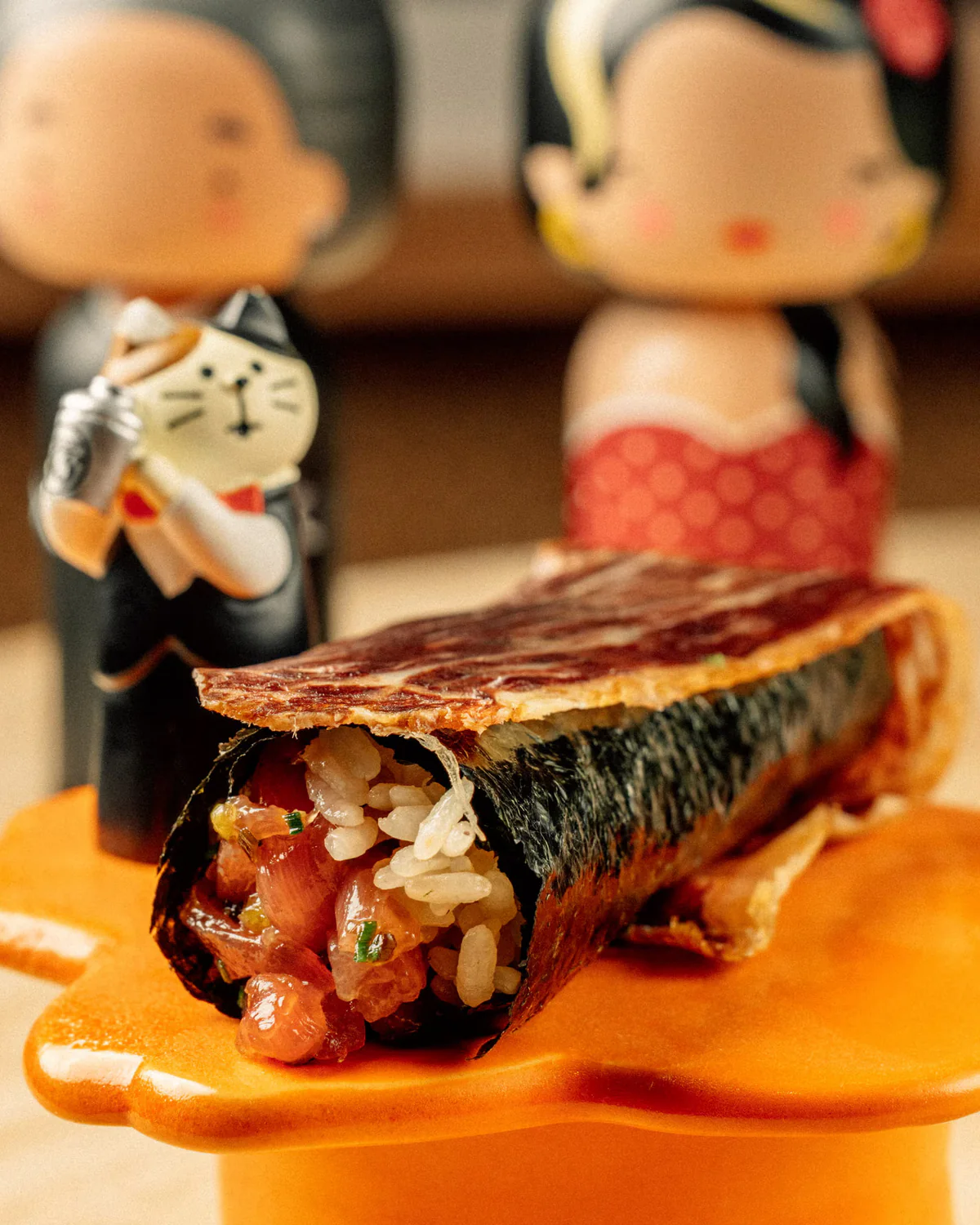 A handroll of chopped tuna and rice topped with a seared slice of wagyu, kokeshi dolls behind